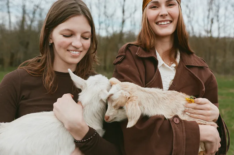 Two cheerful women hold a white goat kid in a sunny countryside setting, sharing a gentle animal moment, casual style, and a relaxed lifestyle scene, while smiles brighten the peaceful outdoor space
