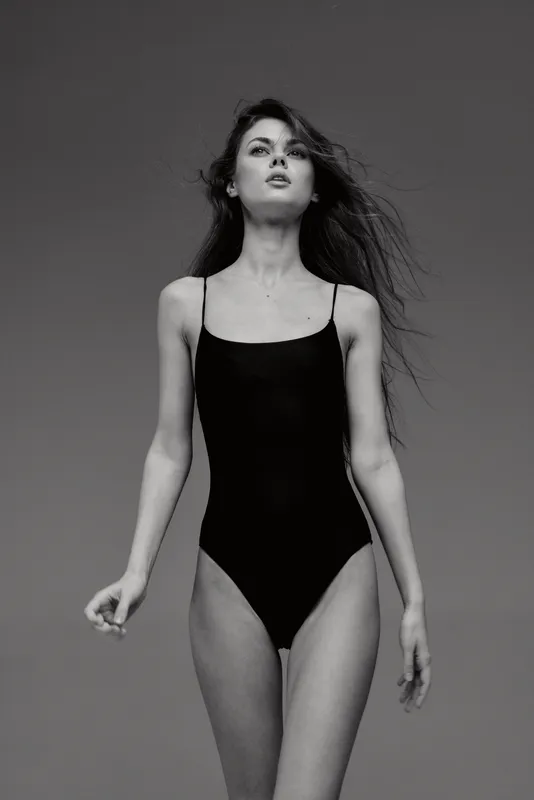 Black and white portrait of a young woman in a black swimsuit with flowing hair looking confident against a dark background.