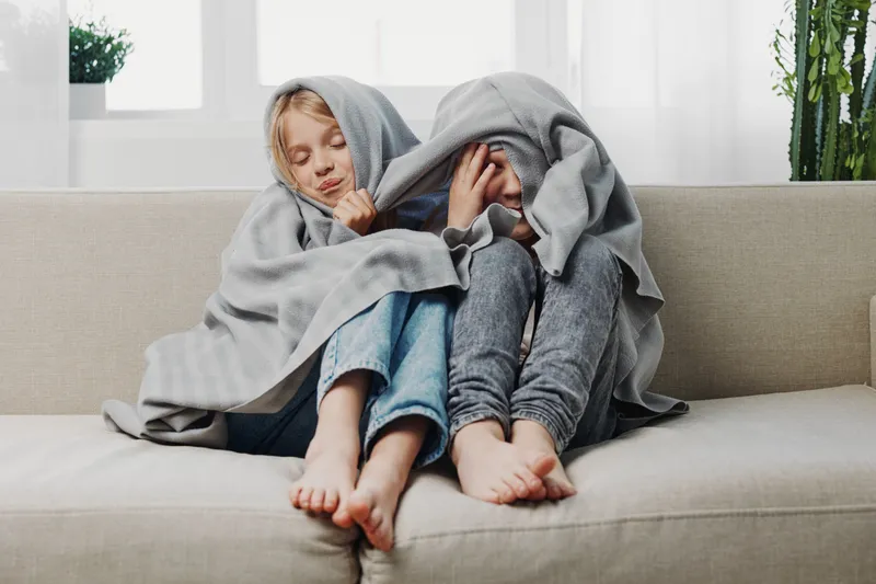 Two children wrapped in grey blankets sit barefoot on a beige sofa in a bright room. The scene captures warmth, comfort, and sibling bonding during a cozy indoor moment.