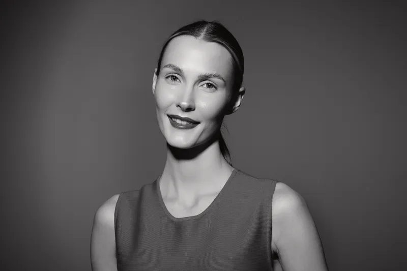 Portrait of a smiling Caucasian woman with light skin and pulled back hair in a sleeveless top on a dark background.