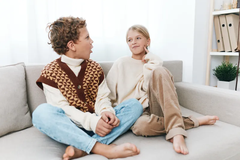 Two children sit close on a cozy living room sofa, wearing warm knit sweaters and casual jeans, sharing a calm, candid moment of connection.
