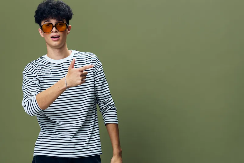 young man in striped shirt with sunglasses pointing finger sideways against olive green background, casual style, confident gesture, studio portrait, positive expression