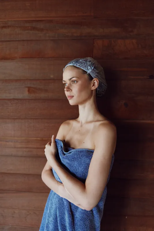woman wrapped in blue towel with shower cap standing against wooden wall, natural light, calm expression, indoor bathroom setting, wellness and self-care concept.