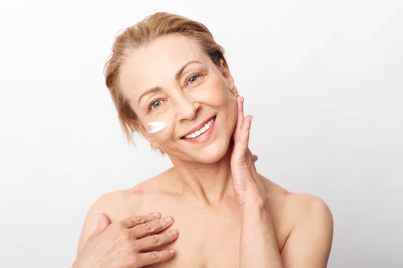 Mature woman applying moisturizing cream on face and shoulder smiles gently. Skincare concept focused on natural beauty, skin health, and self-care routine on white background.