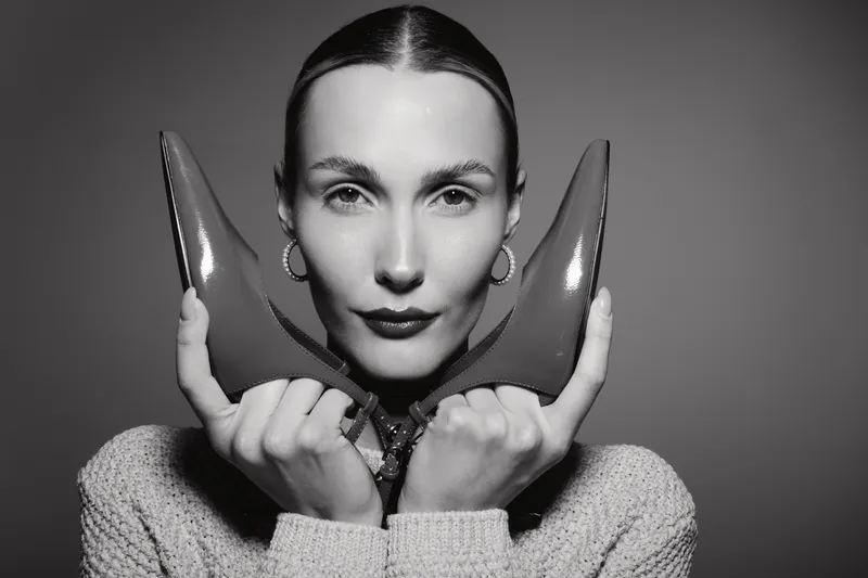 Young white woman with fair skin holds pair of high heel shoes near face, wearing cozy sweater and hoop earrings in studio portrait. Concept of fashion and style.