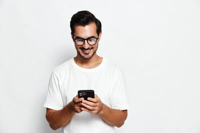 Smiling happy cheerful young man wearing glasses holding a mobile phone in hand, looking at the screen, isolated on plain white background. Modern lifestyle concept.