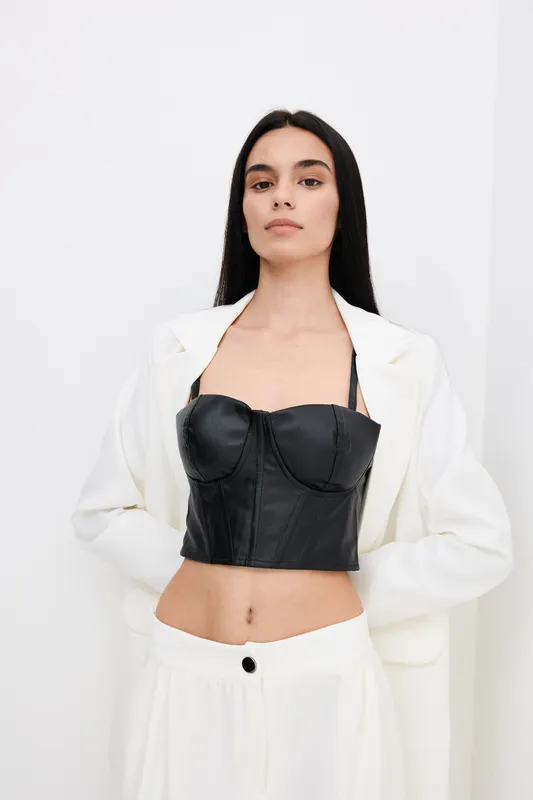 Young woman wearing a black leather bustier and white suit, standing confidently against a plain white background in a contemporary fashion style.