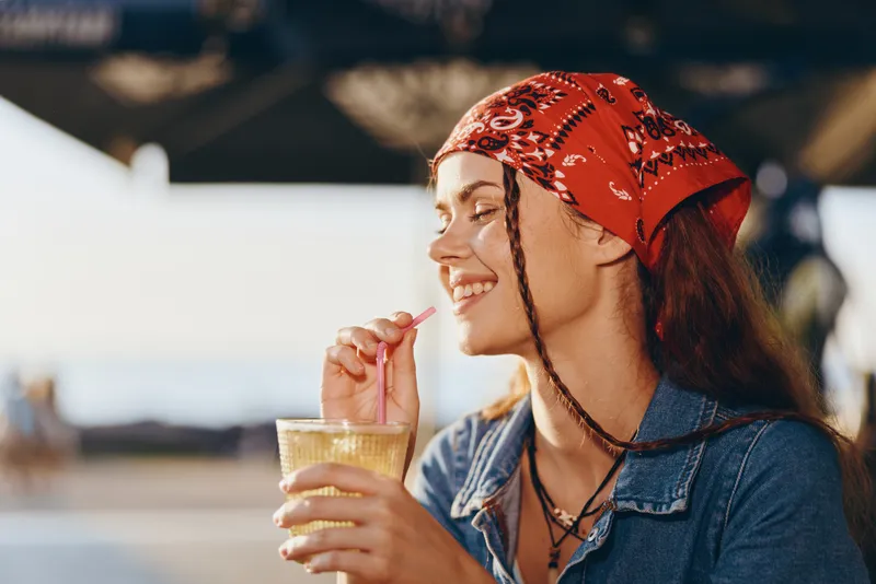 smiling woman drinking cocktail outdoors wearing red bandana and denim jacket relaxed in warm sunlight during summer day