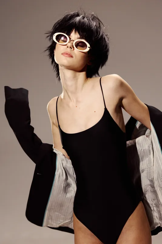 Stylish woman in black swimsuit and sunglasses with short hair posing with jacket in minimal studio setting.