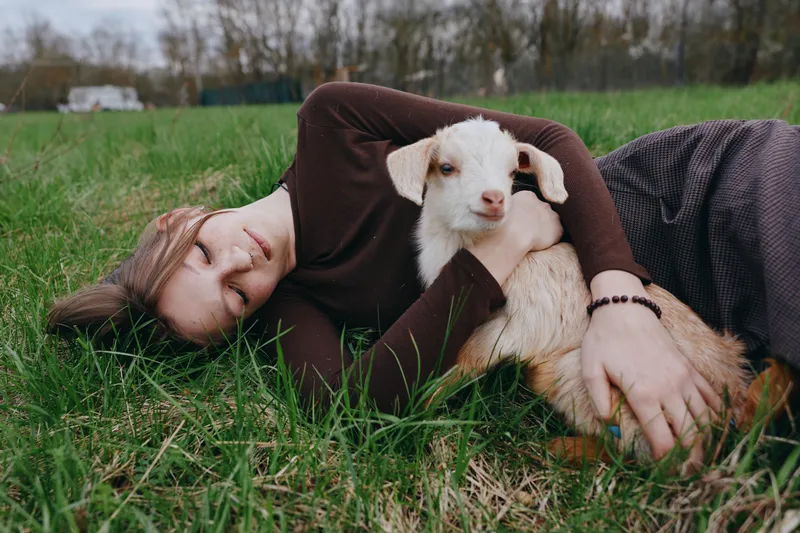 Relaxed woman lying in grass beside a young goat kid, outdoor lifestyle scene, gentle companionship, countryside atmosphere, soft natural light, peaceful mood, candid moment, authentic connection,
