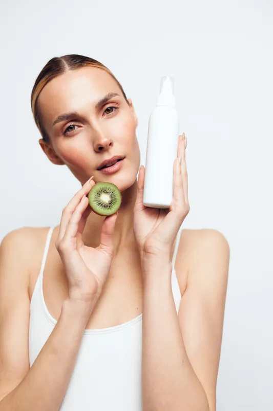 Woman holding skincare serum bottle with kiwi slice, close up on face, clean white studio background, concept of skincare, wellness, self care, soft studio lighting, fresh beauty