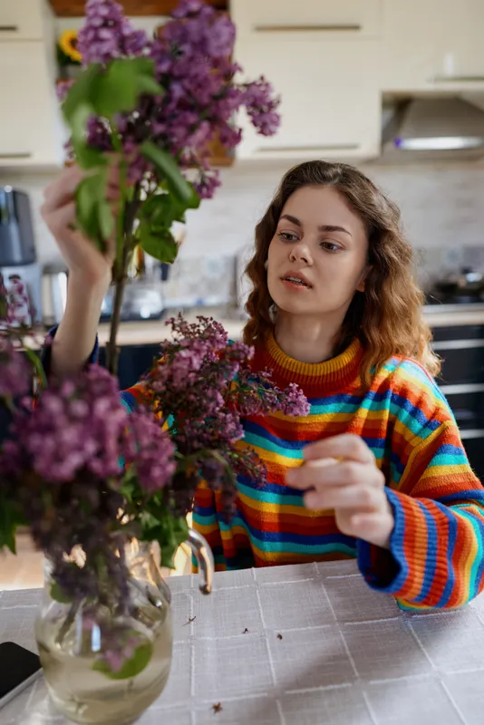A woman in a colorful striped sweater arranges a purple bouquet on a kitchen table, focused and calm, capturing a home lifestyle moment, indoors creativity and flower care, concentration.