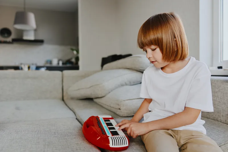 A focused boy playing a red keyboard in a cozy living room, showcasing creativity and enthusiasm while enjoying music with his playful expression.