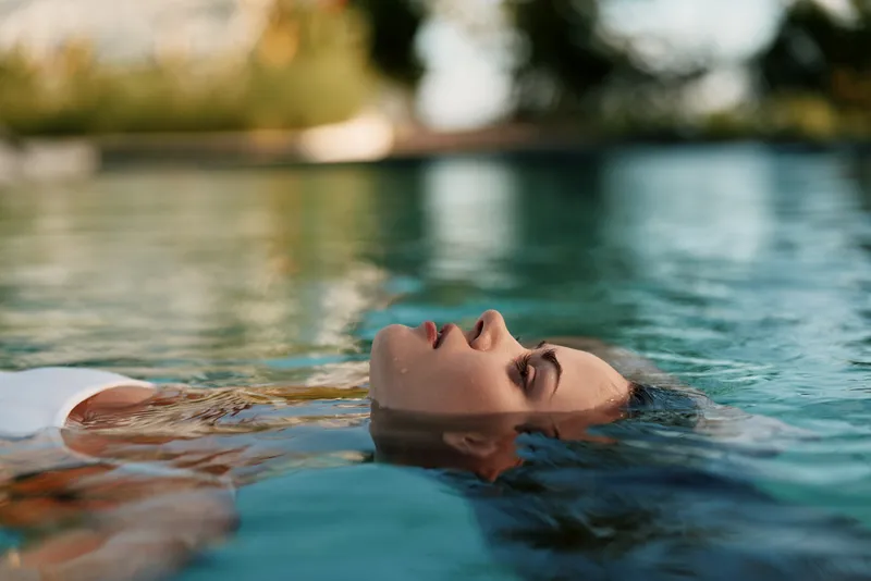 Calm woman floating in a sunlit pool, tranquil outdoor moment with clear water and gentle ripples, relaxed posture, natural colors, serene ambiance, lifestyle scene