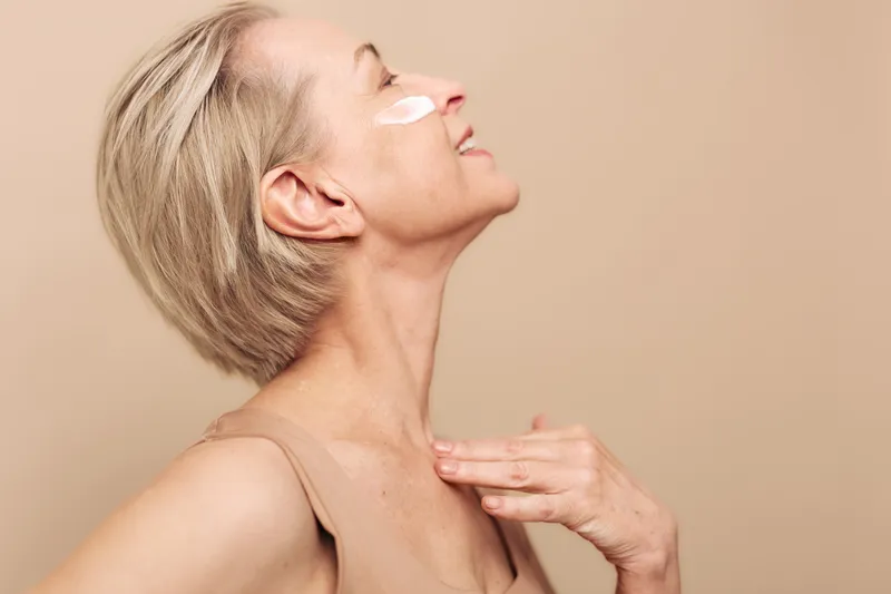 Mature woman with short blond hair applies cream on cheek and chest, smiling with closed eyes on beige background. Concept of skincare, self-care, natural beauty, and healthy lifestyle.