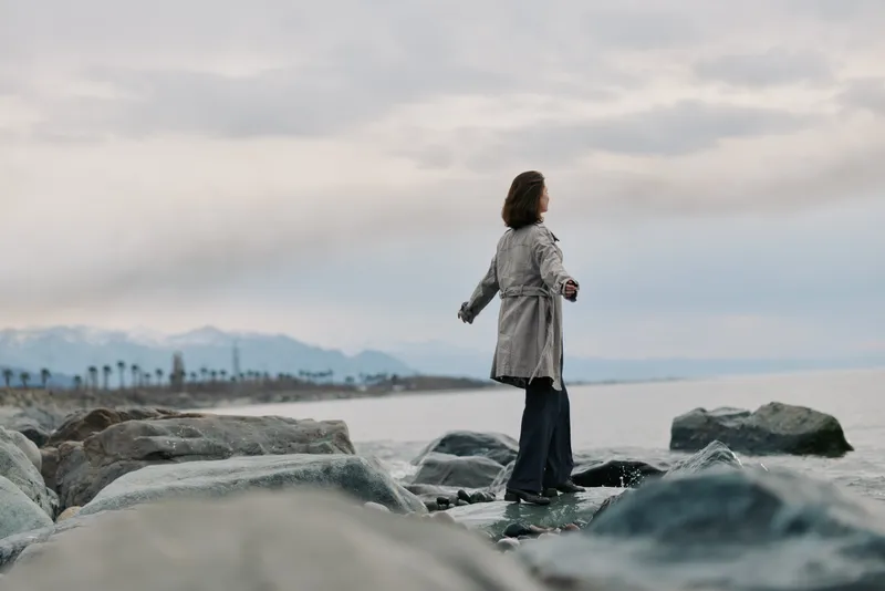 Woman in trench coat stands on rocky shore by the sea under cloudy sky. Calm water and distant mountains create a peaceful outdoor atmosphere and a sense of solitude.