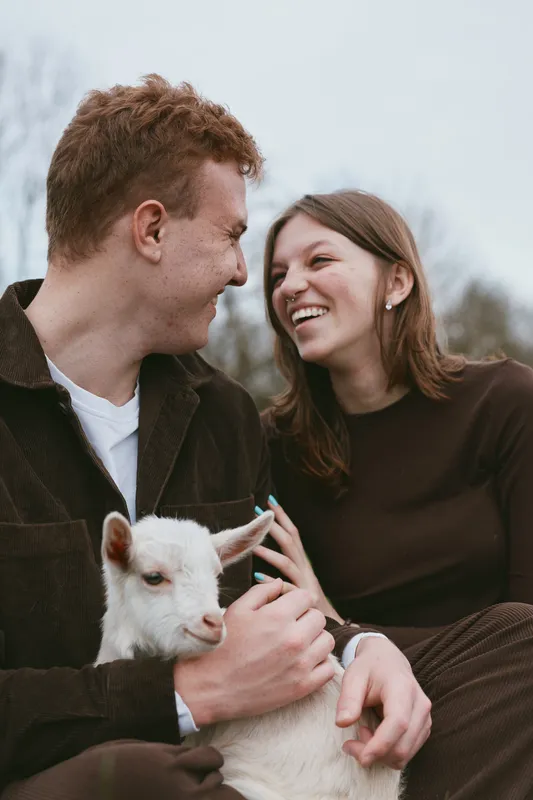 Happy couple outdoors with a goat kid, sharing warm moment and genuine affection, showcasing a relaxed lifestyle, joyful bonding in natural light, candid smiles, rural setting.