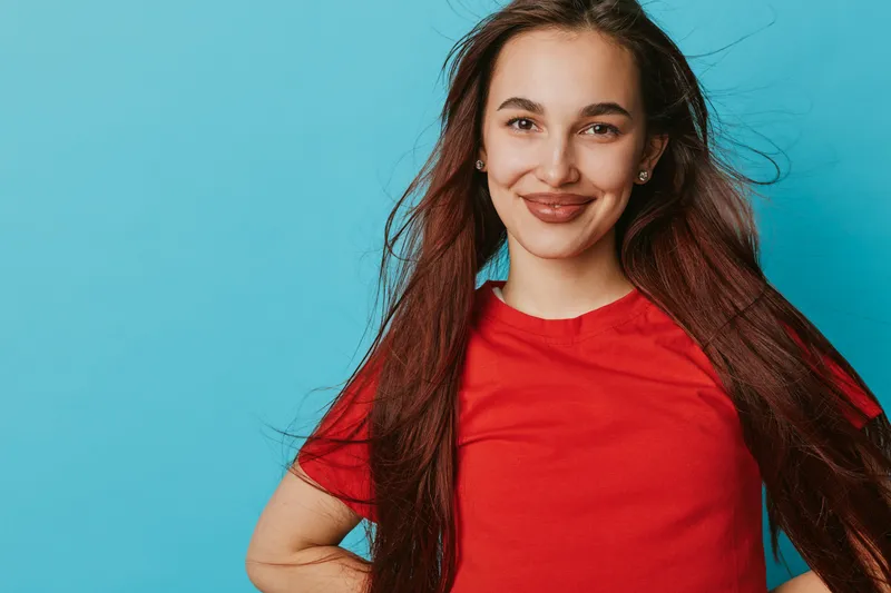 Young woman with long brown hair wearing red t shirt smiling confidently with hand on hip against blue background. Portrait of happy female with natural makeup and earrings.