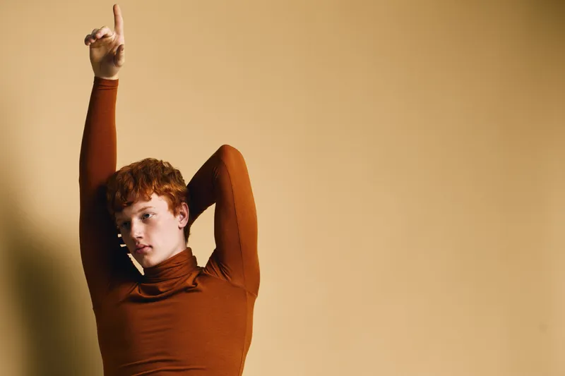 Young man with curly red hair wearing brown turtleneck posing with arm up against beige background in studio. Fashion, portrait and style with confident expression.