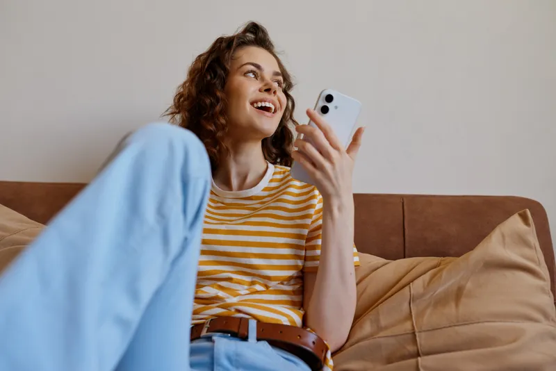 Cheerful woman relaxing on a couch at home, smiling at her smartphone, casual striped tee, soft natural light, cozy living room, lifestyle moment