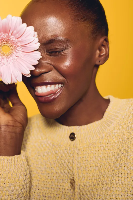 Smiling dark skinned African woman in yellow sweater holds pink flower near face, eyes closed, showing healthy glowing skin. Concept for beauty, skincare, and natural happiness.