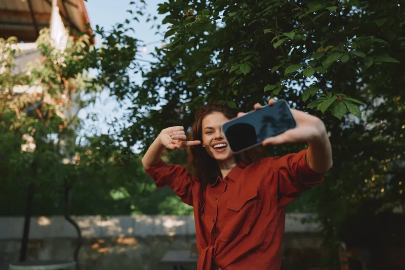Young woman takes a carefree outdoor selfie wearing a red shirt, smiling broadly, capturing a candid lifestyle moment in a sunlit garden setting, vibrant and joyful