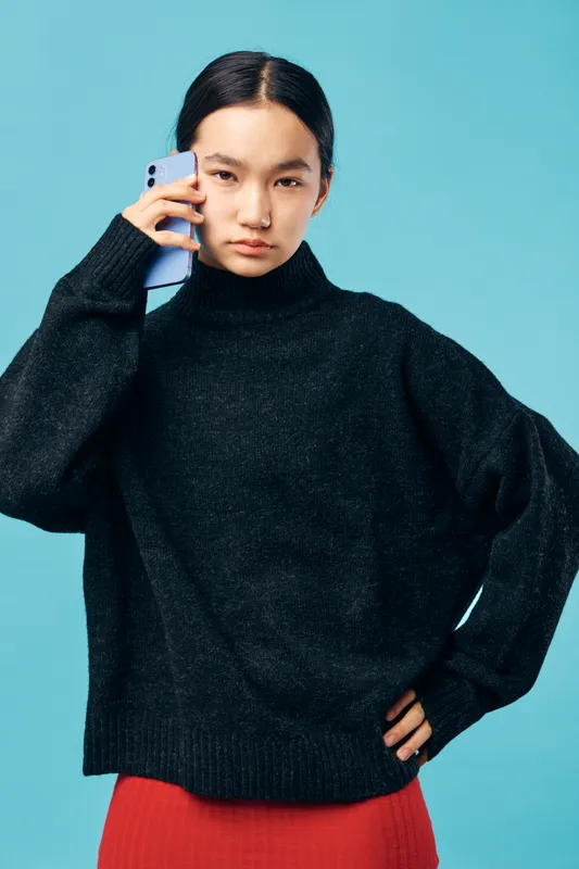 Young woman wearing black sweater with hand on hip talking on smartphone with serious expression against blue background. Casual style, technology and communication concept.