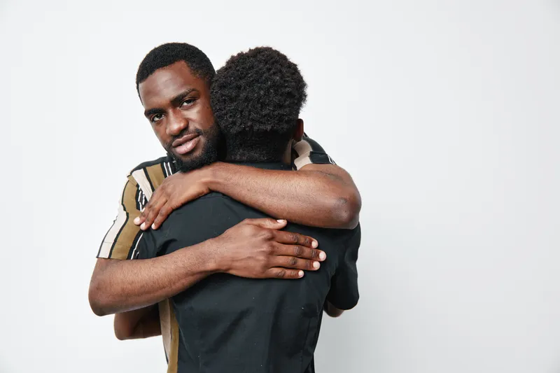 Two black men hugging each other in a warm embrace on white background. African ethnicity, dark skin, casual clothing, emotional connection, friendship and support between friends.