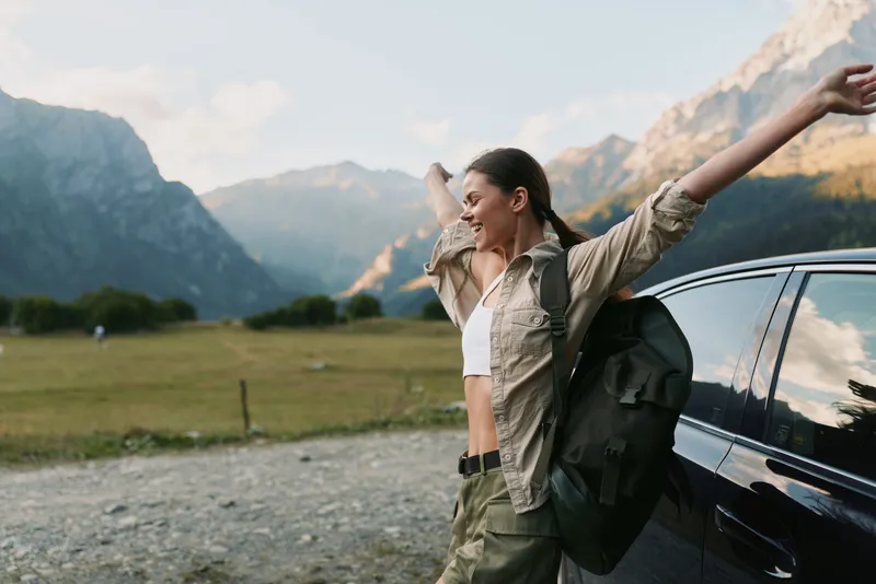 Young woman with a backpack stretches her arms joyfully next to a car in a mountain valley during a sunny day, enjoying freedom and adventure in nature.