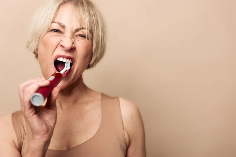 Blonde woman brushing teeth with electric toothbrush against beige background. Close-up of oral hygiene routine and dental care concept with focused expression and casual outfit.