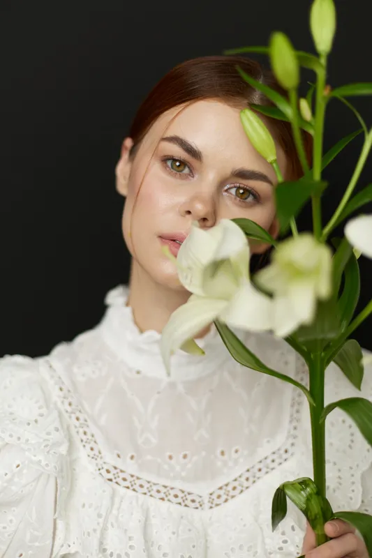 A serene young woman in a white dress holding a white flower against a dark background, conveying beauty, innocence, and natural elegance
