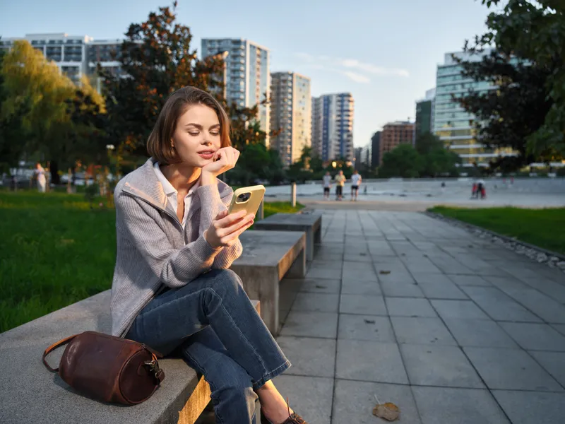 A calm urban professional sits on a bench in a modern city park, looking at a smartphone with a thoughtful smile, surrounded by trees and contemporary buildings.