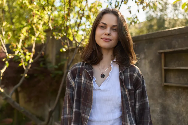 Smiling woman with plaid shirt standing outdoors in warm sunlight, casual lifestyle, natural confidence, and everyday wellness. Bright background, soft shadows