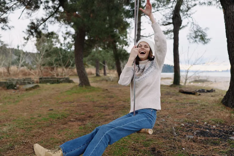 Joyful woman on a swing in a forested park, laughing and carefree, casual sweater and jeans, nature scene, playful moment outdoors