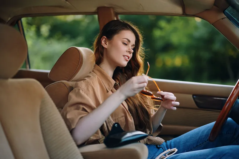 Young woman with long hair wearing a brown shirt, sitting in a car and adjusting her sunglasses in a sunny outdoor setting, expressing a relaxed and carefree mood.