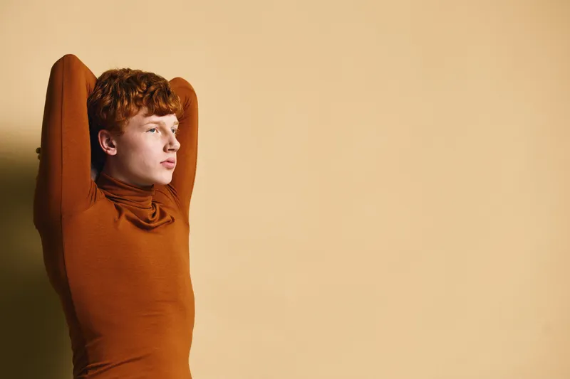 Young man with red hair wearing brown turtleneck stretching arms behind head against beige background. Casual style and thoughtful pose with side view and relaxed mood.