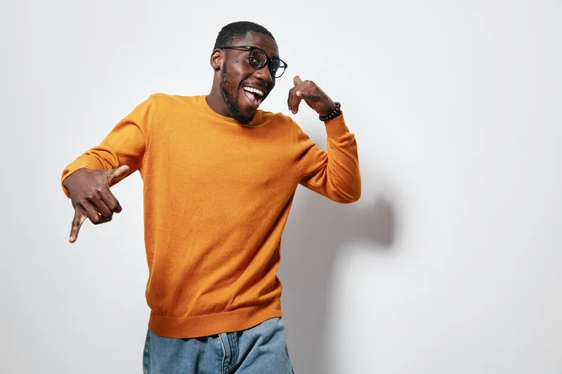 African American man with dark skin wearing orange sweater and glasses posing happily with hand gestures on white background. Casual style and joyful expression in studio.
