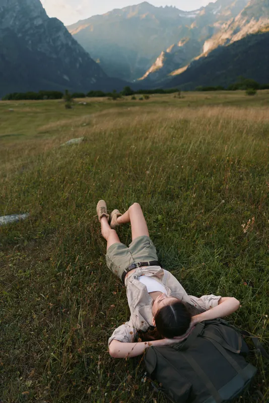 Young woman lies on grass in open field surrounded by mountains enjoys peaceful nature moment with casual outfit and backpack in warm daylight outdoors lifestyle scene