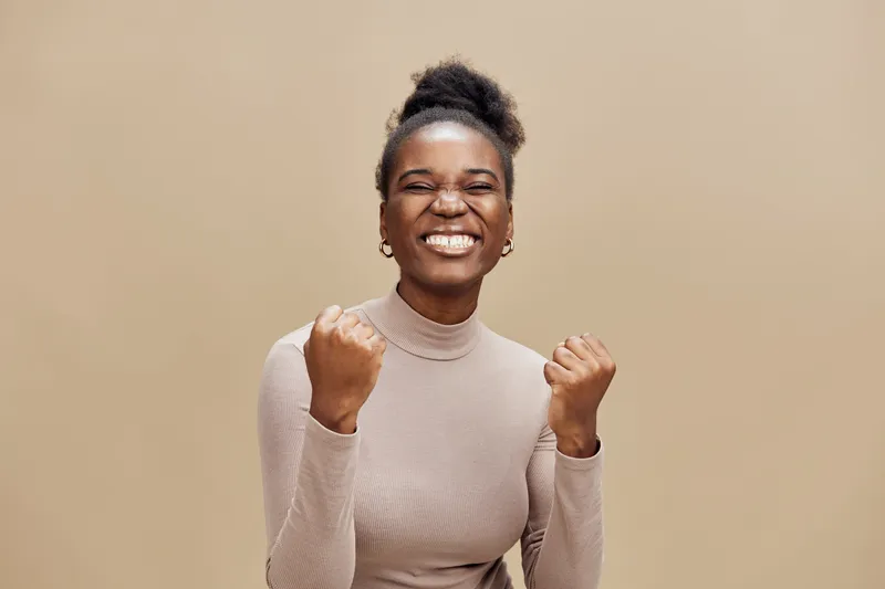 Happy African woman celebrating success in a light beige background, wearing a stylish turtleneck and expressing joy and excitement with clenched fists