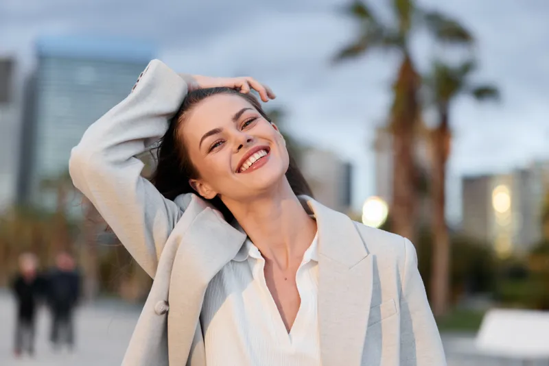 Happy woman smiling outdoors in stylish light coat and white shirt in urban park during daytime with blurred cityscape and palm trees background