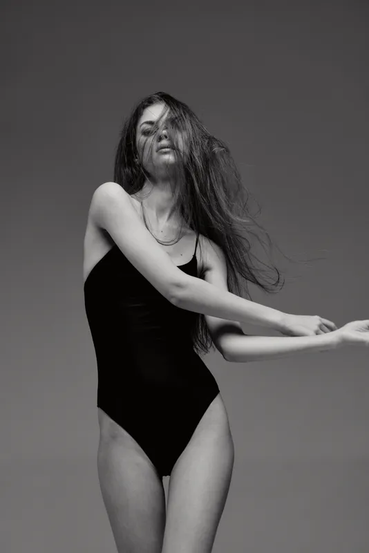 Black and white portrait of a young woman in a black bodysuit with flowing hair, posing gracefully against a plain background.