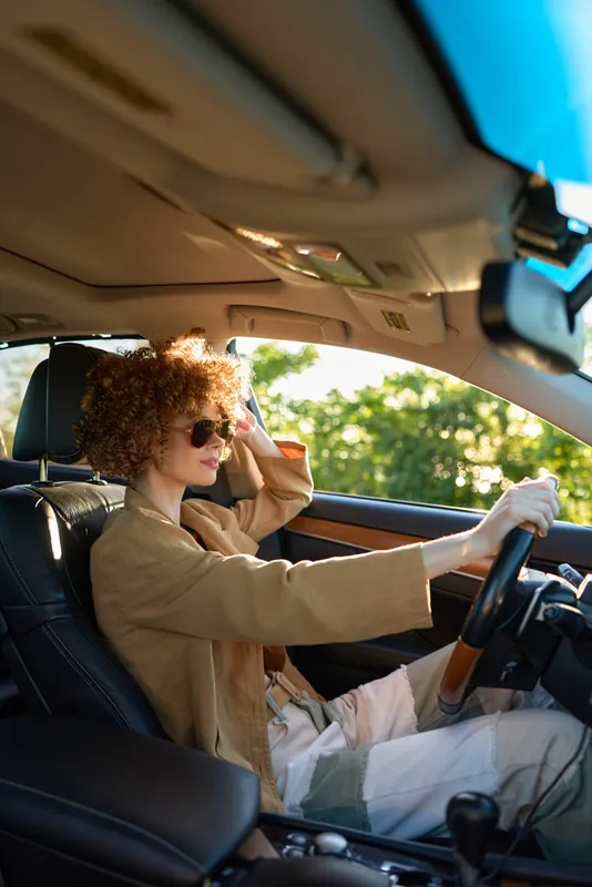Cheerful woman behind the wheel, sunlit car interior, relaxed smile, casual jacket, retro film color palette, warm glow, candid lifestyle moment