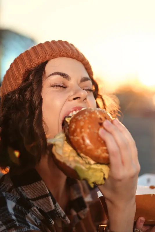 Woman biting a burger in a candid moment, burger bite and smile framed by golden hour glow. Authenticity and mindful living captured in emotional storytelling street food portrait.