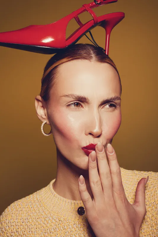 Woman with red high heel shoe balanced on head wears yellow sweater and hoop earrings. She covers mouth with hand and looks sideways on brown background. Concept of fashion and style.