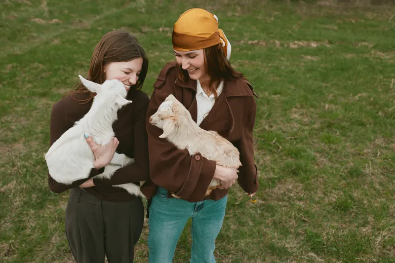 A candid lifestyle scene shows two women outdoors with a playful goat kid, smiles and gentle interaction capturing farm charm, warmth, and connection in a natural setting.