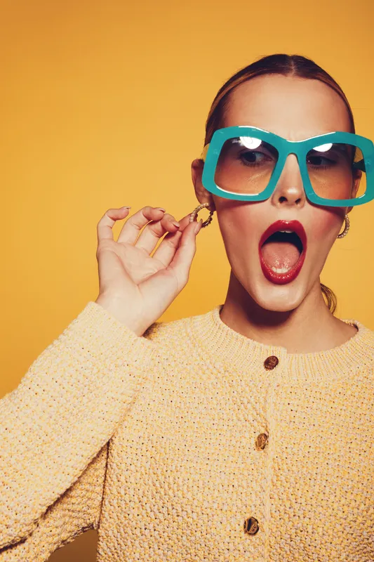 Stylish woman in oversized turquoise sunglasses and yellow knitted sweater poses with open mouth on yellow background. Concept of fashion, retro style, summer accessories, and bold makeup.