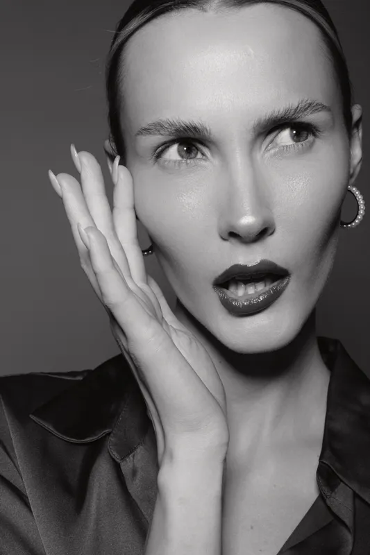 Close-up of a fair skinned white woman with slicked hair and glossy lips posing with hand near face in dramatic studio lighting on black background.