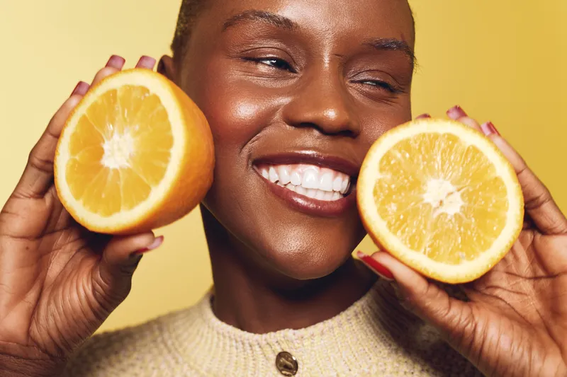 Close-up of a happy black woman with dark skin holding two orange halves near her face on a yellow background. Concept of healthy lifestyle, vitamin C, and natural beauty.