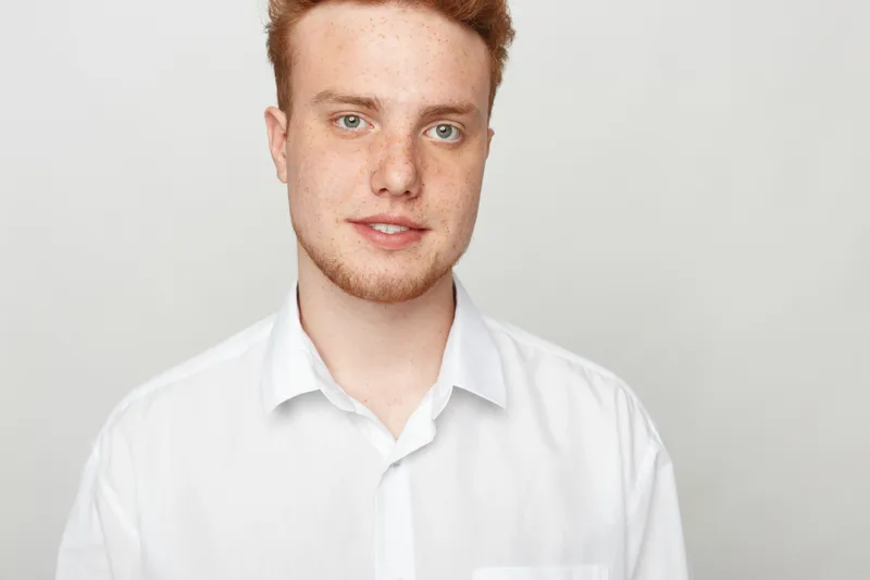 Portrait of a young man with red hair and beard wearing a white shirt against a plain light background. Casual style, confident expression, natural lighting, studio shot.
