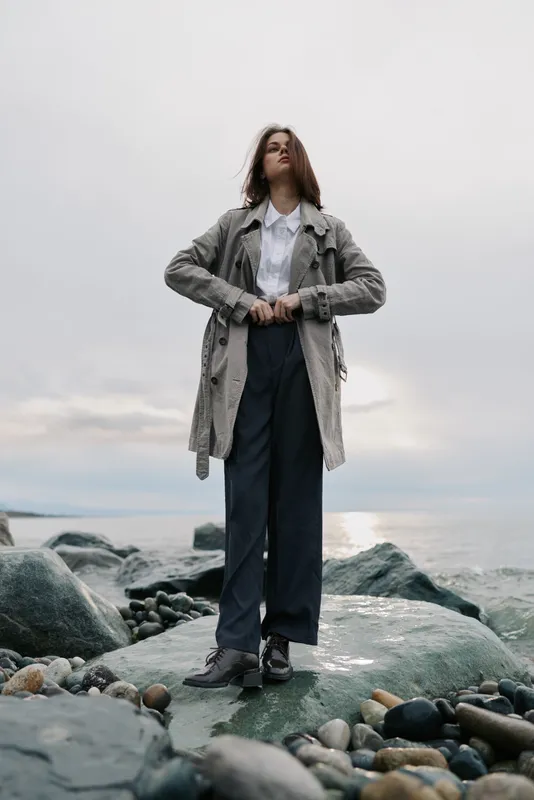 Tall woman in a grey coat and white shirt stands on rocky shore under cloudy sky. Fashion concept with confident pose and natural outdoor background near water. Moody atmosphere.